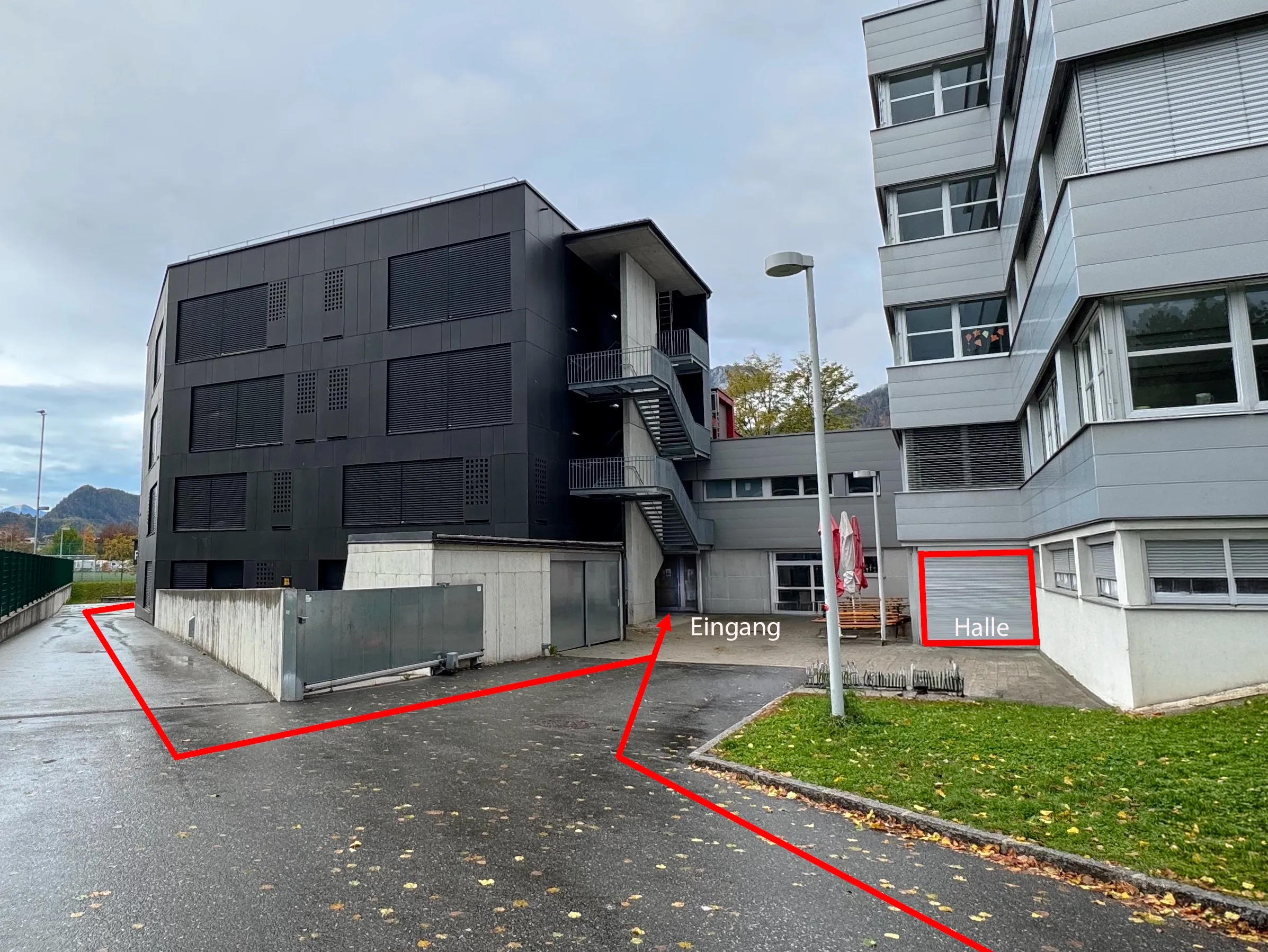 Weg zur Trainingshalle an der Mädchen-HS Kufstein: Rote Linien zeigen die Wege von links entlang des dunklen Gebäudes und von vorne über den Vorplatz zum Eingang zwischen den beiden Gebäuden, rechts ist die Halle markiert.